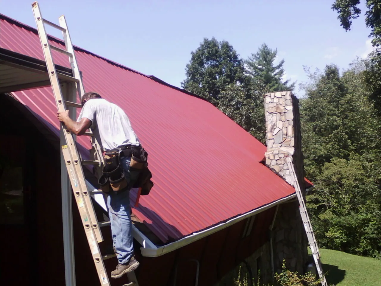 Roofer on a ladder beside a red metal roof and stone chimney — Soffit & Fascia Repair by Roofing Brothers in Julian